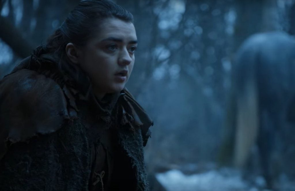 Arya sits at a campfire, looking to the right of the frame in the snow