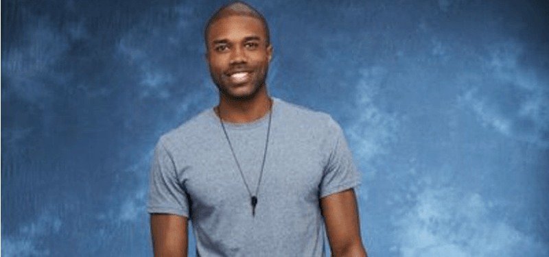 DeMario is wearing a grey shirt and is standing in front of a blue background.