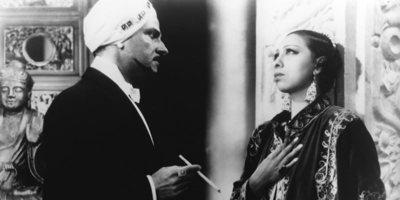 Josphine Baker has her hand over her chest as a man looks at her holding a cigarette.