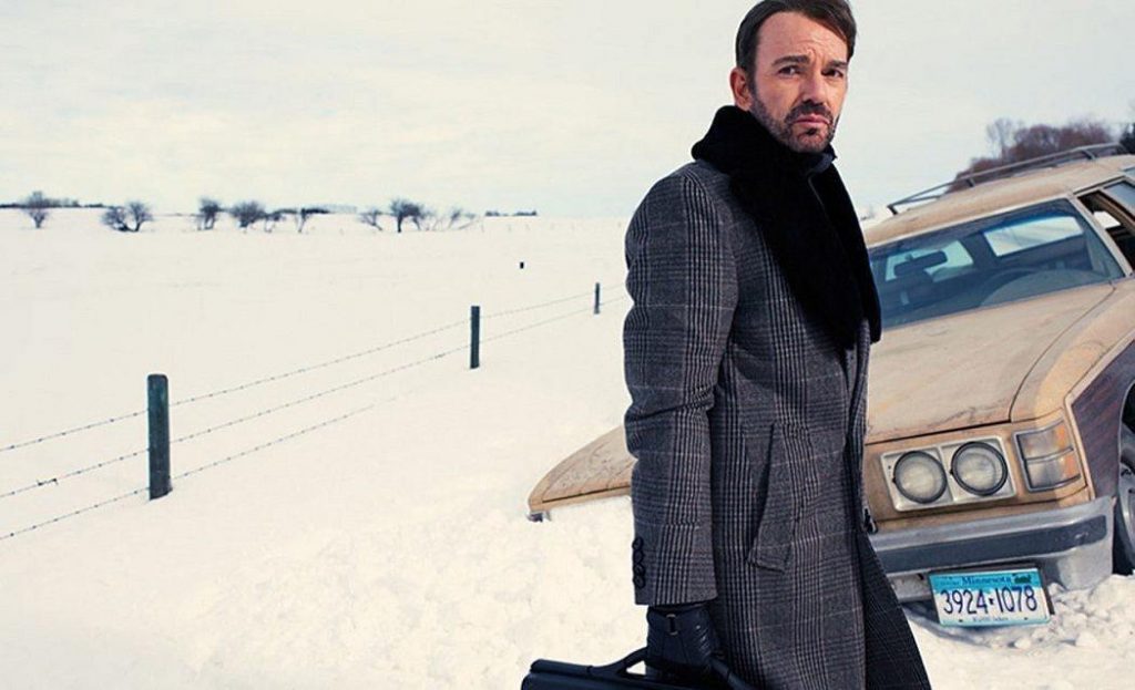 Billy Bob Thornton as Lorne Malvo, wearing a long grey trenchoat in the snow, holding a suitcase in his right hand