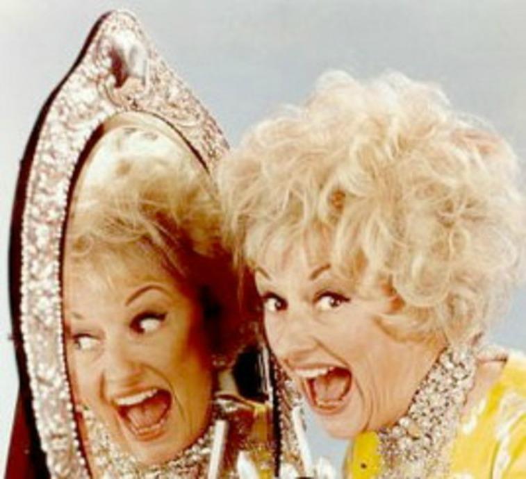 Phyllis Diller sonriendo con la boca abierta junto a un espejo donde se ve su reflejo