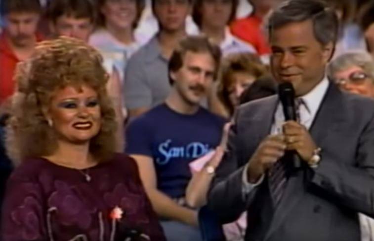 Tammy Faye está de pie junto a su marido Jim Bakker, que sostiene un micrófono frente a la audiencia del estudio's holding a microphone in front of a studio audience