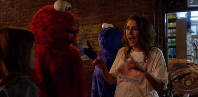 Gretchen has messed up hair and is wearing a t-shirt will standing next to Elmo on the street.
