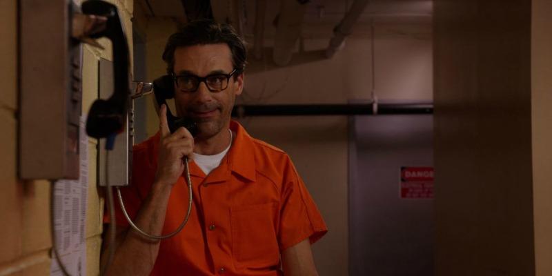 Jon Hamm is in an orange jumpsuit and is talking at a payphone in prison.