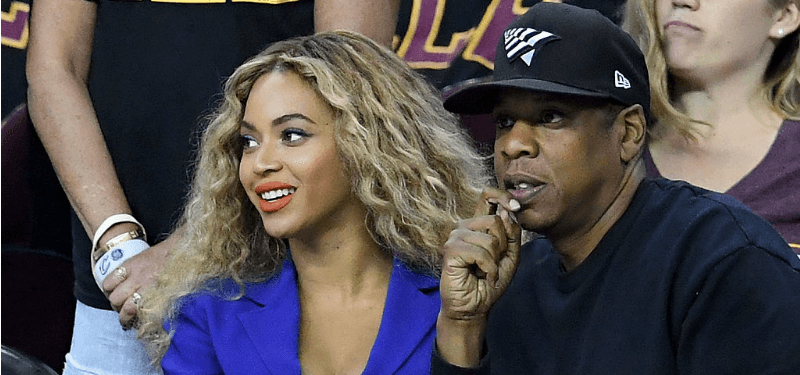 Beyonce is in a blue suit and Jay are watching a NBA Finals game.