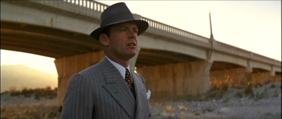 Jack Nicholas, standing under a bridge, wearing a fedora, and looking off into the distance