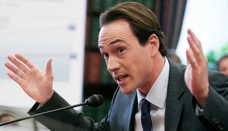 Chris Klein is talking into a microphone with two hands up in the air.