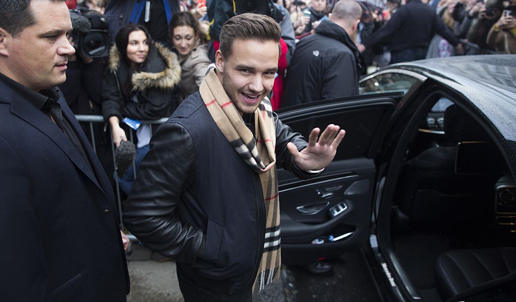 Liam Payne smiling and waving, while getting into a black car and wearing a scarf