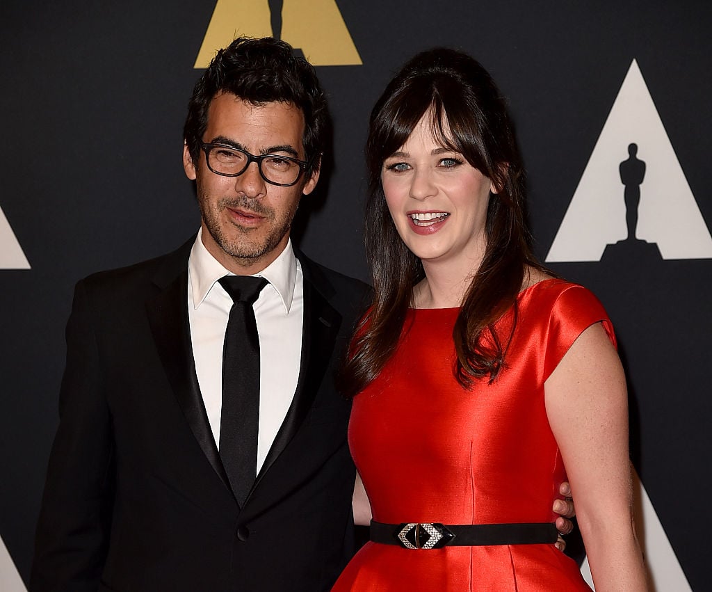 Zooey Deschanel in a red dress, smiling for the camera with her husband Jacob Pechenik on the red carpet