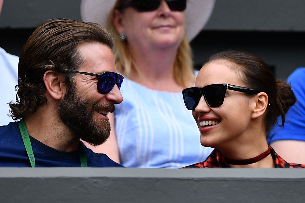 Bradley Cooper and Irina Shayk smiling at each other wearing sunglasses, sitting in the crowd at Wimbledon together