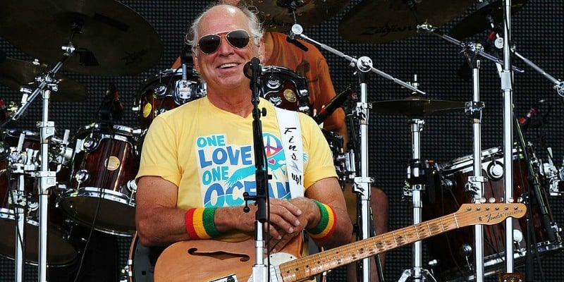 Jimmy Buffett is smiling and resting his hands on his guitar while on stage.