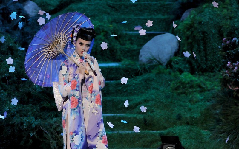 Katy Perry is dressed as a geisha and is holding an umbrella while singing on stage.