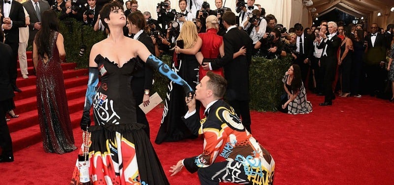 Katy Perry and Jeremy Scott are in matching dress and suit jacket as he is one bended knee kissing her gloved hand.