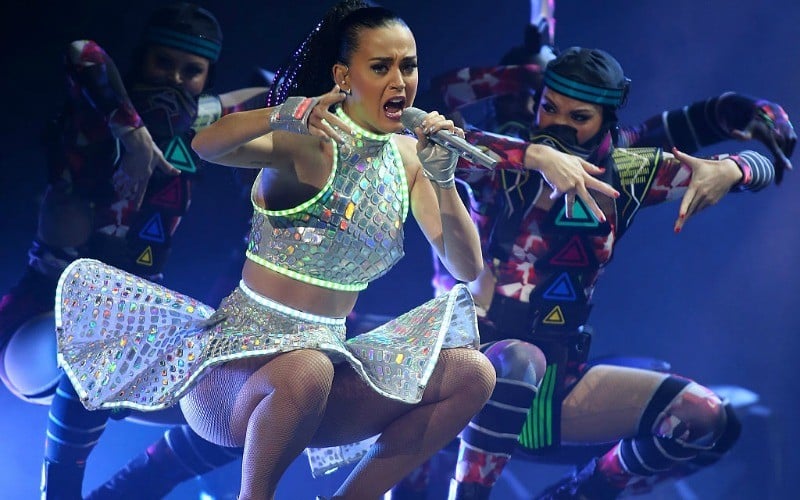 Katy Perry is in a sparkly top and skirt as she is bending down singing on stage.