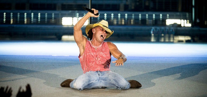 Kenny Chesney is on the ground performing on stage and is holding a microphone in the air.