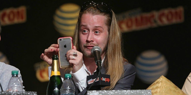Macaulay Culkin is sitting on a panel and is taking a picture with his phone.