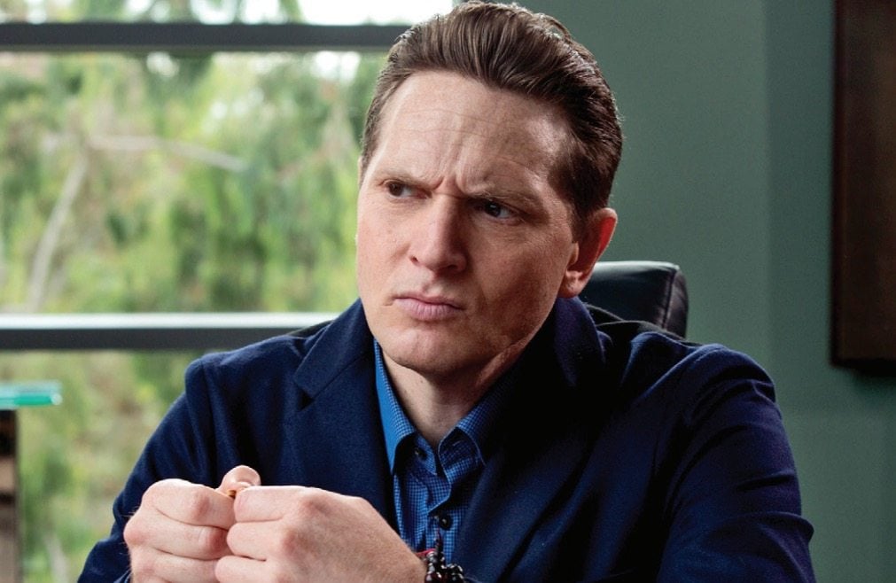 Gavin Belson in a blue sweater, with his hands folded, and looking thoughtfully off to his right