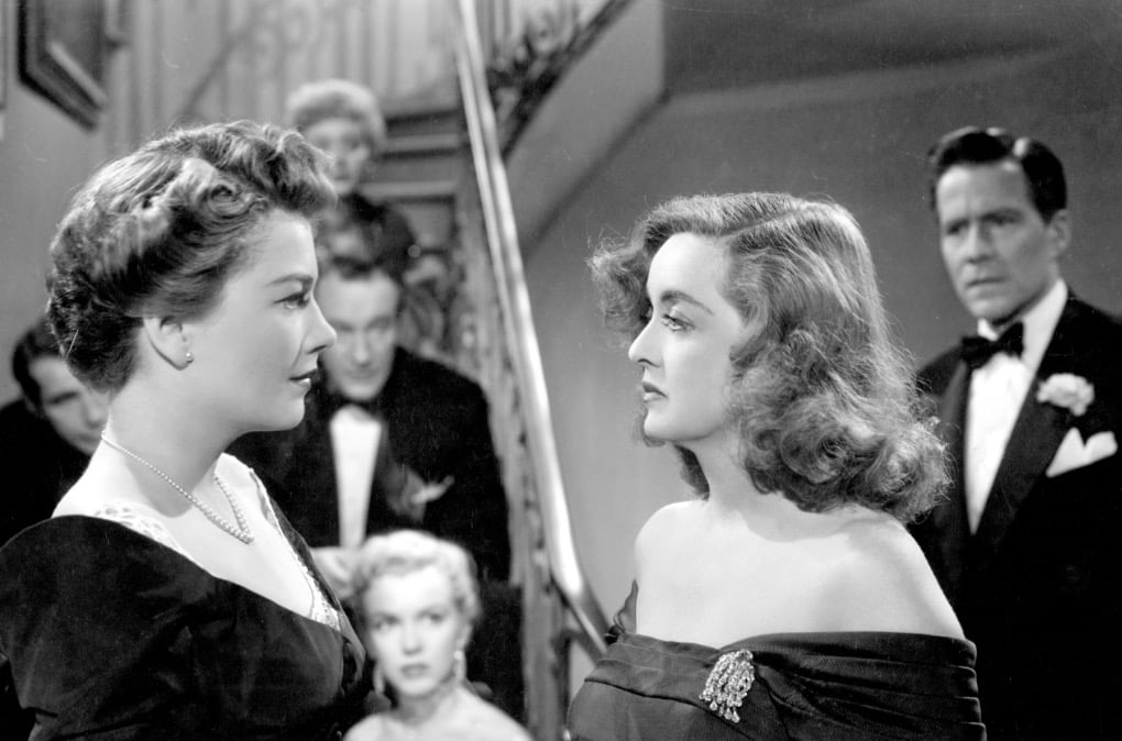 Bette Davis and Anne Baxter, staring intently at each other in front of a crowd of people