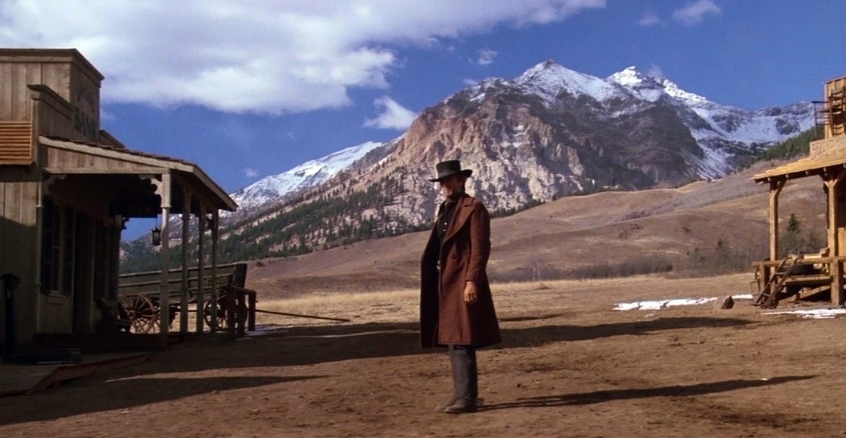 Clint Eastwood in Pale Rider, standing in the middle of a deserted Western town square, with a mountain far in the background