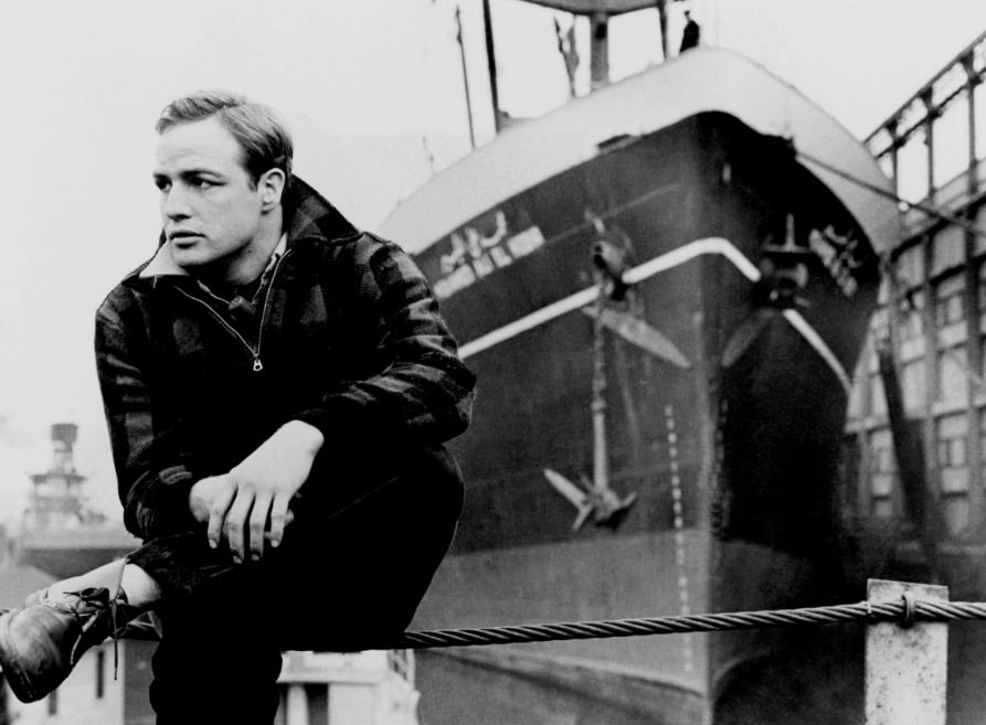 Marlon Brando crouching next to a ship on a dock, looking off over his right shoulder