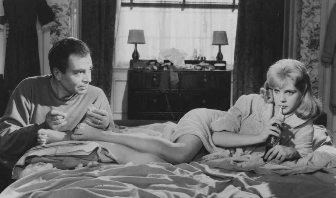 A woman lays on her side on a bed, with a man sitting at the foot of the bed looking at her