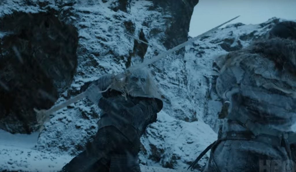 A White Walker swings a sword at a man crouched low to the ground, as the two fight in the snow in front of a ridge