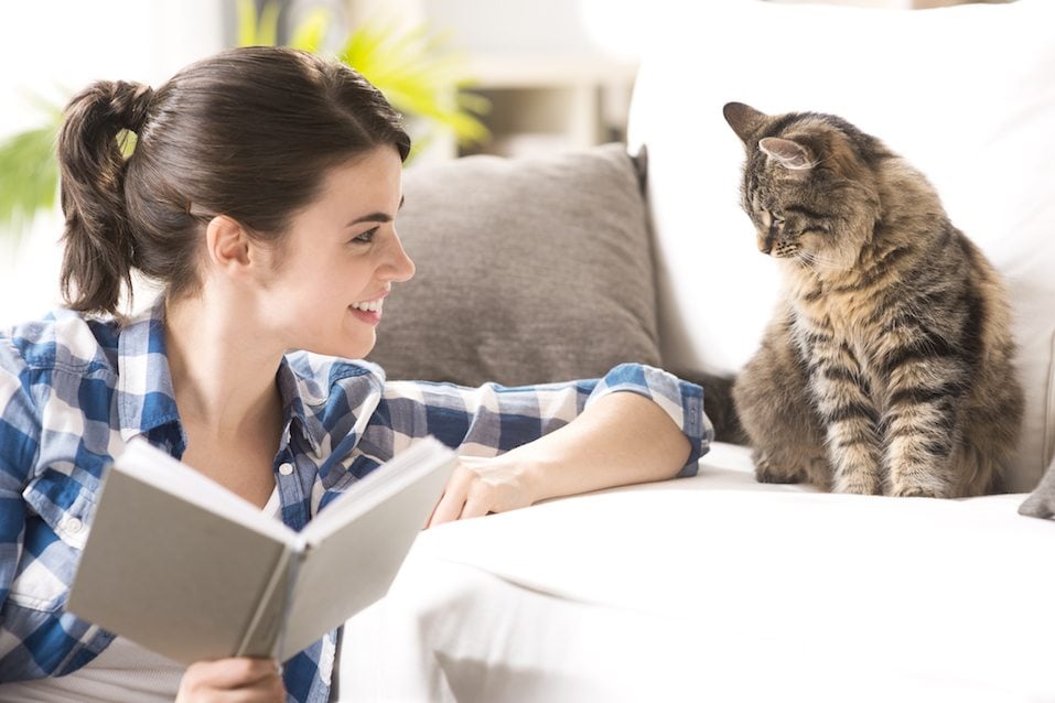 Smiling woman playing with her cat and holding a book in the living room.
