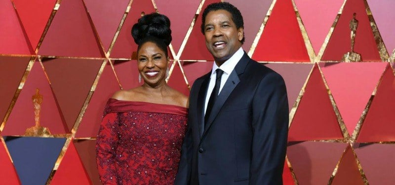 Denzel Washington is in a black suit with Pauletta who is in a red dress.