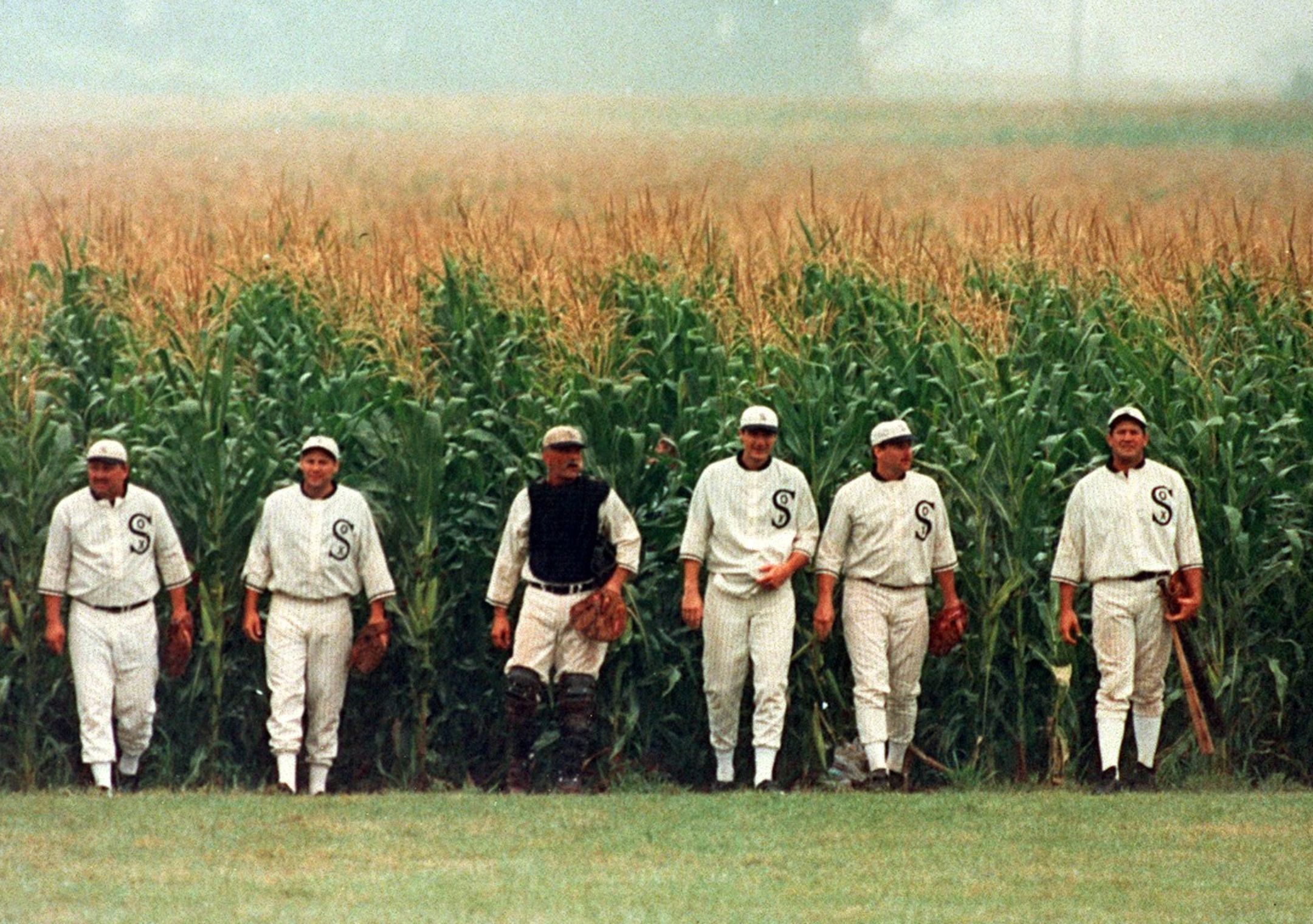 Six men dressed in old baseball uniforms, walking toward the camera out of a corn field