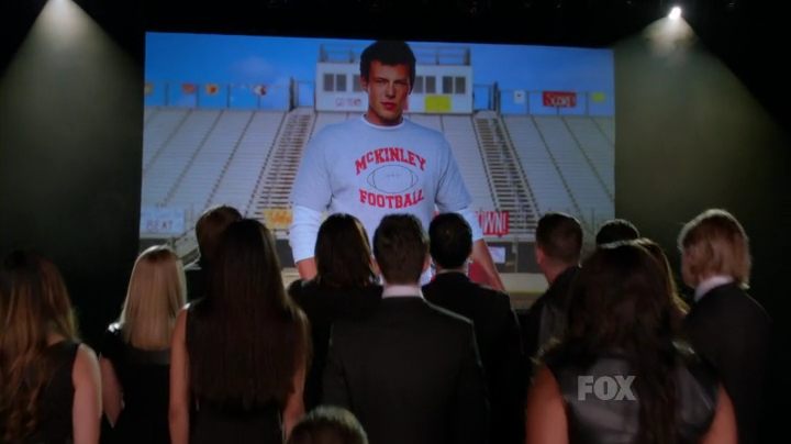 A group of people dressed in back facing away looking at a projected image of a young man posing in a stadium