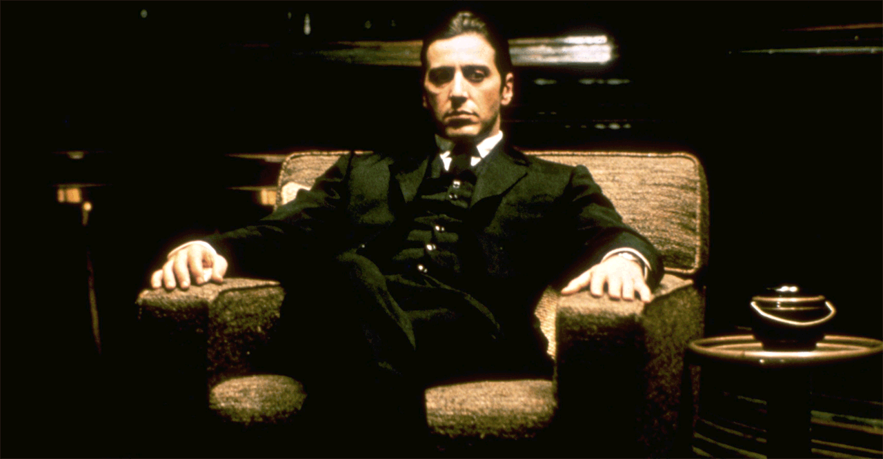 Al Pacino as Michael Corleone, sitting sternly in an armchair and wearing a suit