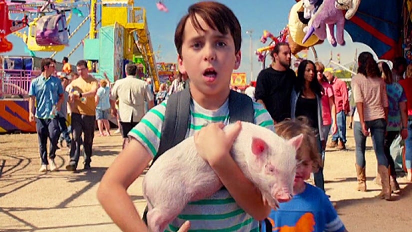 Jason Drucker holds a pig while standing in an outdoor carnival in Diary of a Wimpy Kid: The Long Haul