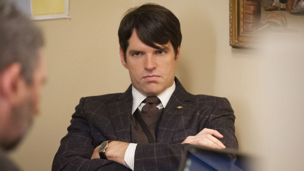 Jonah Ryan wearing a suit, scowling, and crossing his arms defiantly.