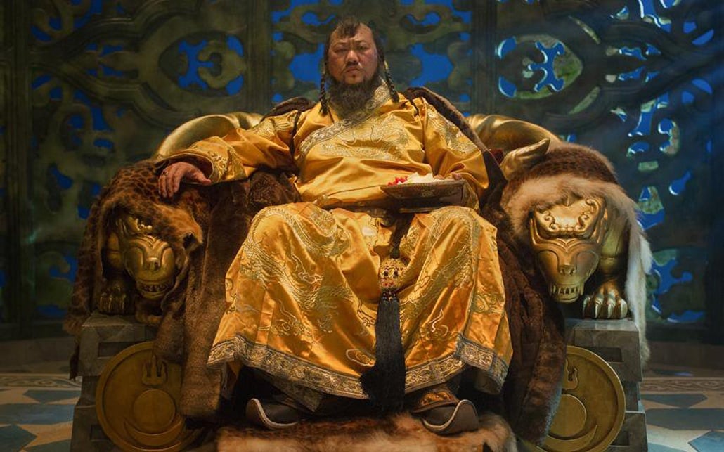 Kublai Khan dressed in a silk yellow robe, sitting atop his thrown with a furrowed brow