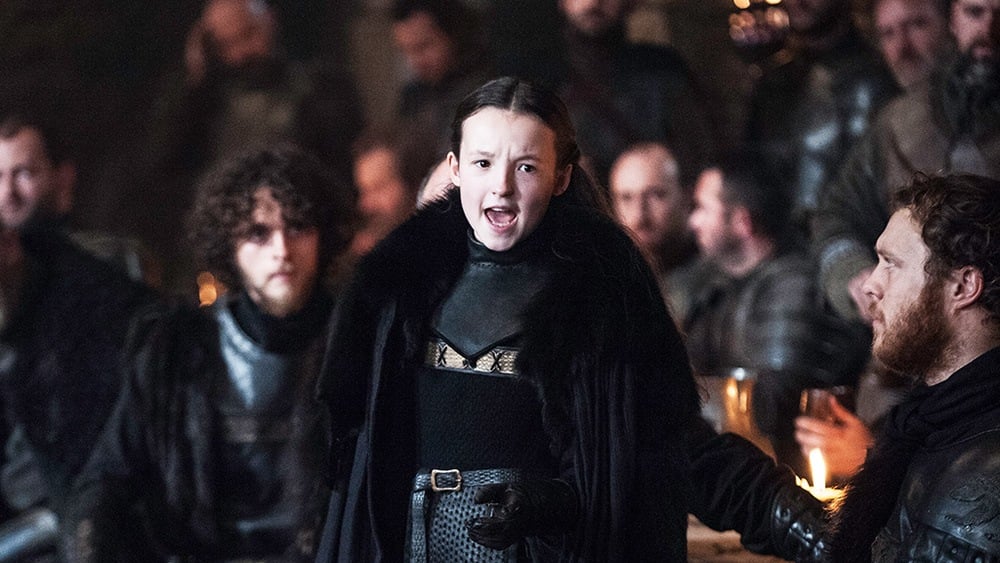 Lyanna Mormont wearing a large black fur coat, speaking to a hall full of men in Winterfell