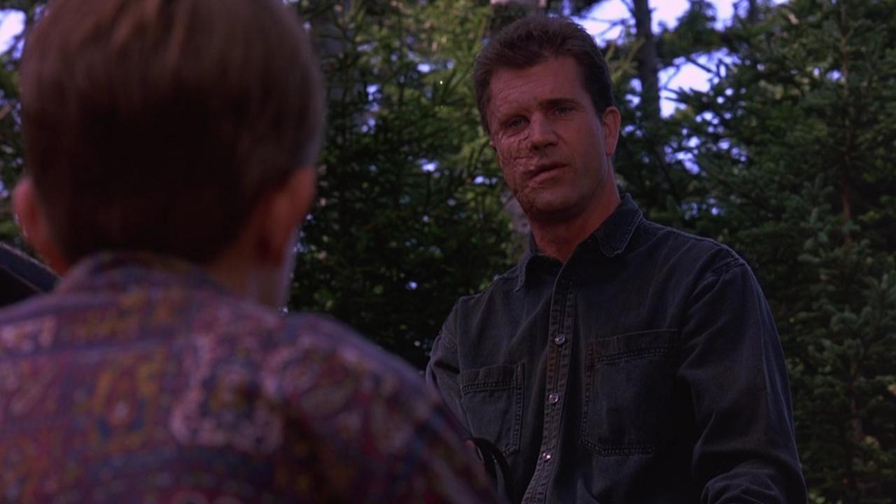 A disfigured Mel Gibson speaks to a small boy in a lightly wooded area