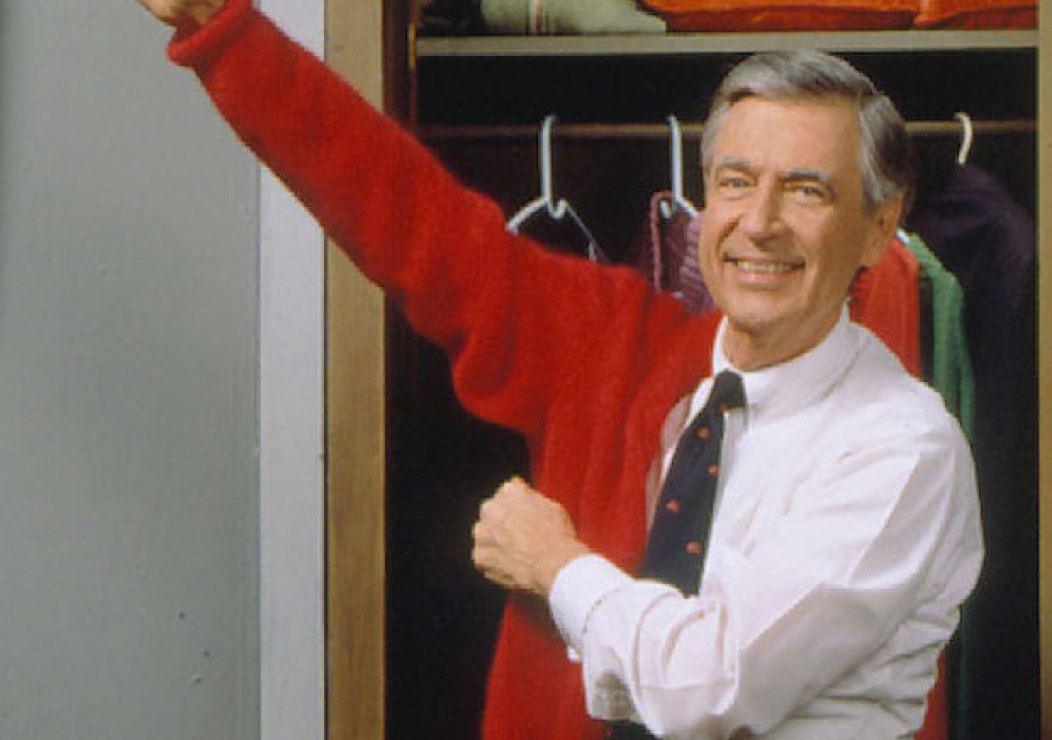 Fred Rogers de pie frente a un armario abierto poniéndose un suéter rojo
