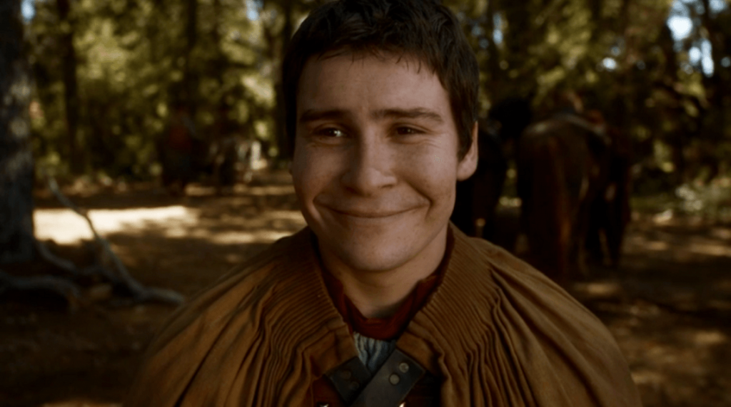 Podrick, wearing a simple brown tunic, and smiling widely into the camera