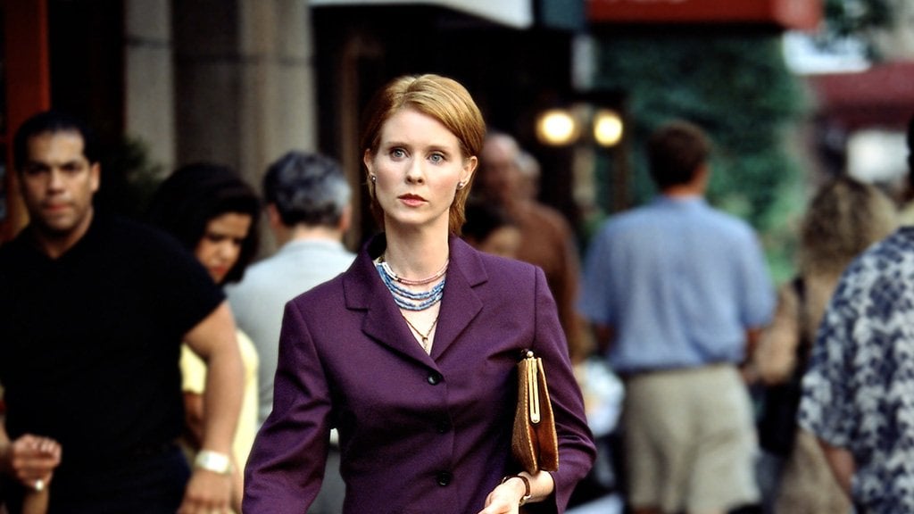 Miranda in a purple blazer with a purse under her arm walks through a crowd of people