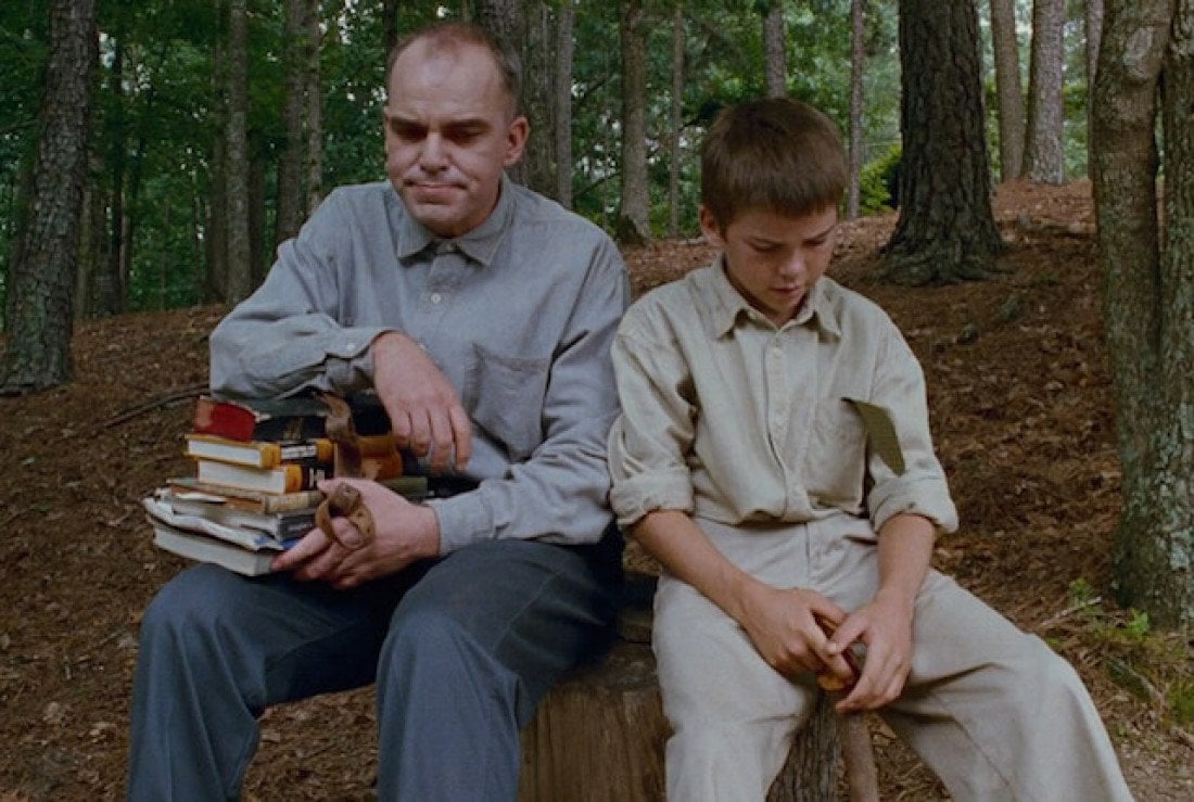 Billy Bob Thornton, sitting on a stump next a child, while resting his arm on a stack of books
