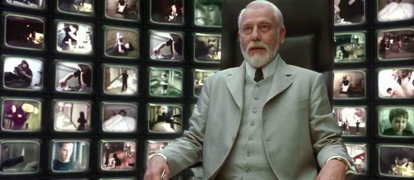 The Architect, wearing a white suit, sitting in front of a large bank of TVs playing in the background