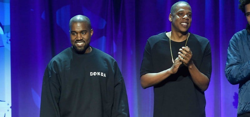 Jay Z is clapping and standing next to Kanye West.