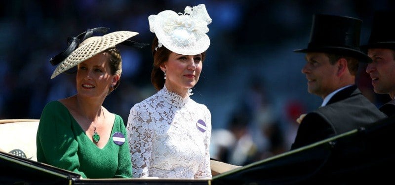 Kate Middleton is sitting in a carriage in a white lace dress and hat.