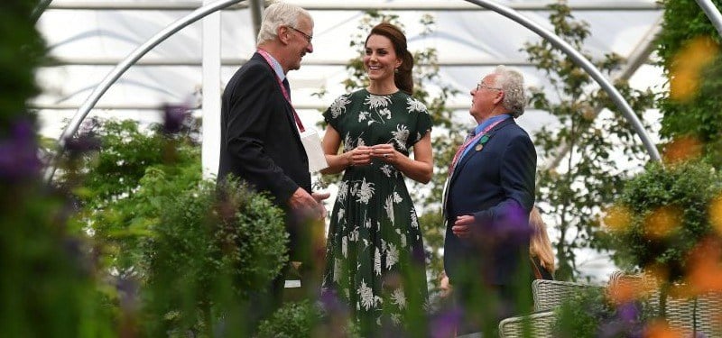 Kate Middleton is in a green floral dress and talking to two men in a greenhouse. Kate Middleton is in a green floral dress and talking to two men in a greenhouse.