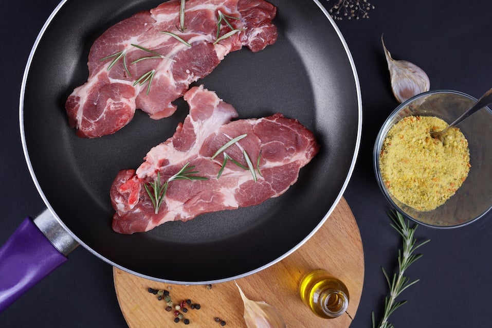 Raw pork neck meat in nonstick frying pan, spices, vegetables, olive oil.