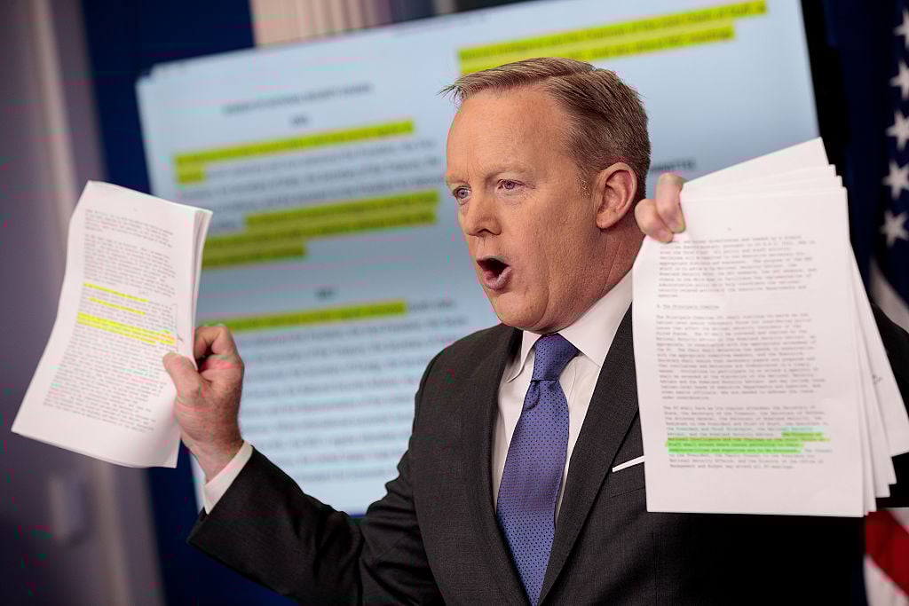 Sean Spicer is talking and holding up two packets of paper. Sean Spicer is talking and holding up two packets of paper.
