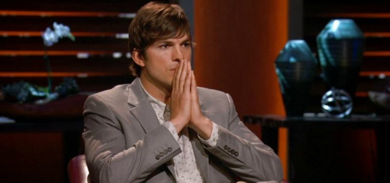 Ashton Kutcker is sitting in a grey suit and has his hands together in front of his mouth.