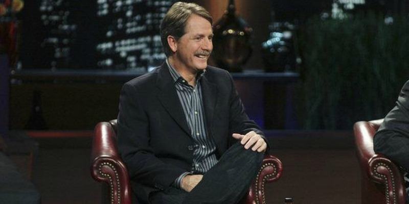 Jeff Foxworthy is sitting in a suit on Shark Tank smiling.