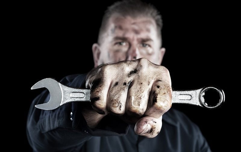 A mechanic covered in grime and grease holds out a box wrench during repairs.