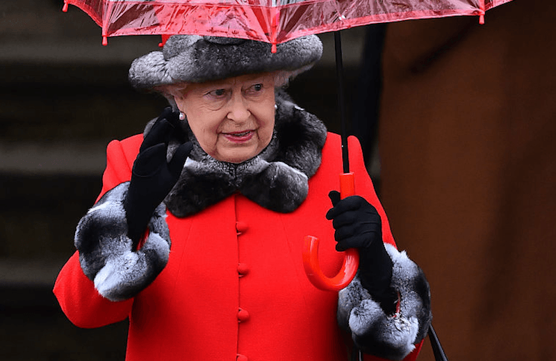 The Queen walks with an umbrella and a red jacket with fur accents.